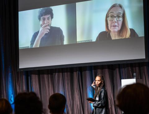 Die Moderatorin spricht vor den an die Leinwand geworfenen Videokacheln der Keynote-Speaker.