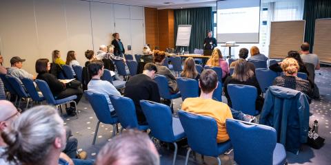 Teilnehmende sitzen in Stuhlreihen in einem Workshop vor einem Referenten.