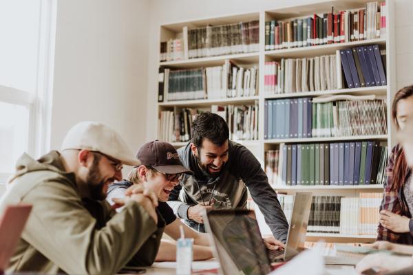 Lachende junge Menschen schauen gemeinsam auf einen Bildschirm im Hintergrund ein Bücherregal