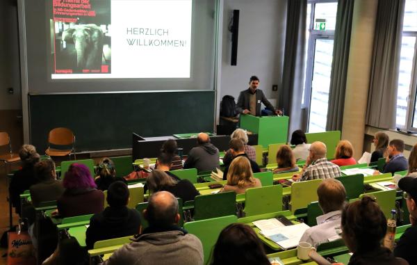 Hörsaal mit Referent vor Pult und Screen mit Teilnehmenden