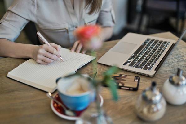 Person an Tisch mit Notizbuch, Laptop, Handy und einer Tasse Kaffee