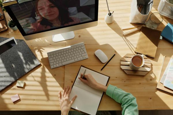 Schreibtisch mit Bildschirm, Tastatur, Kaffeetasse und Person mit Stift und Schreibblock